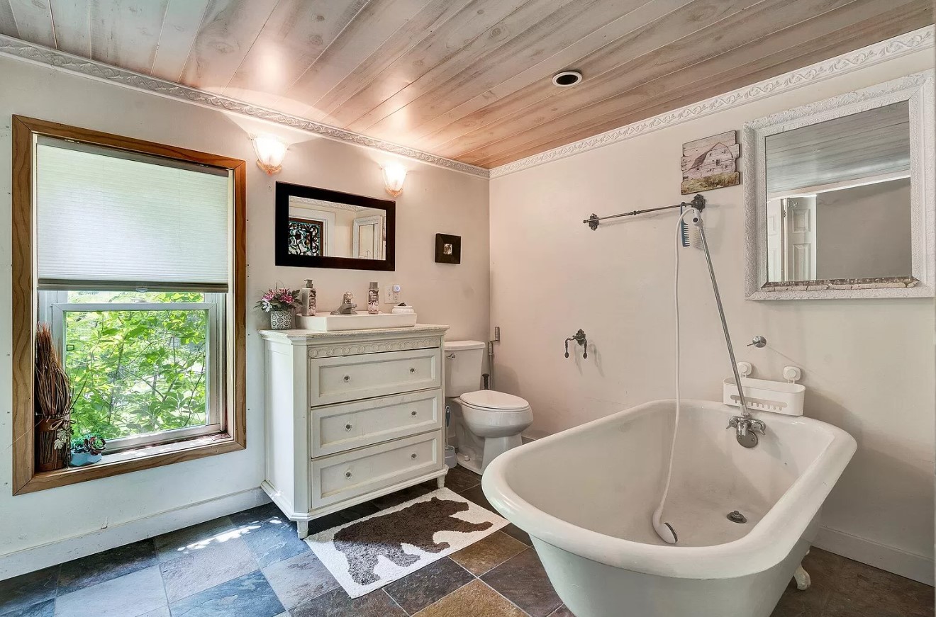 bathroom of an old cabin, white big bath in the middle, white cabinet with three drawers and a mirror above, big window next to it