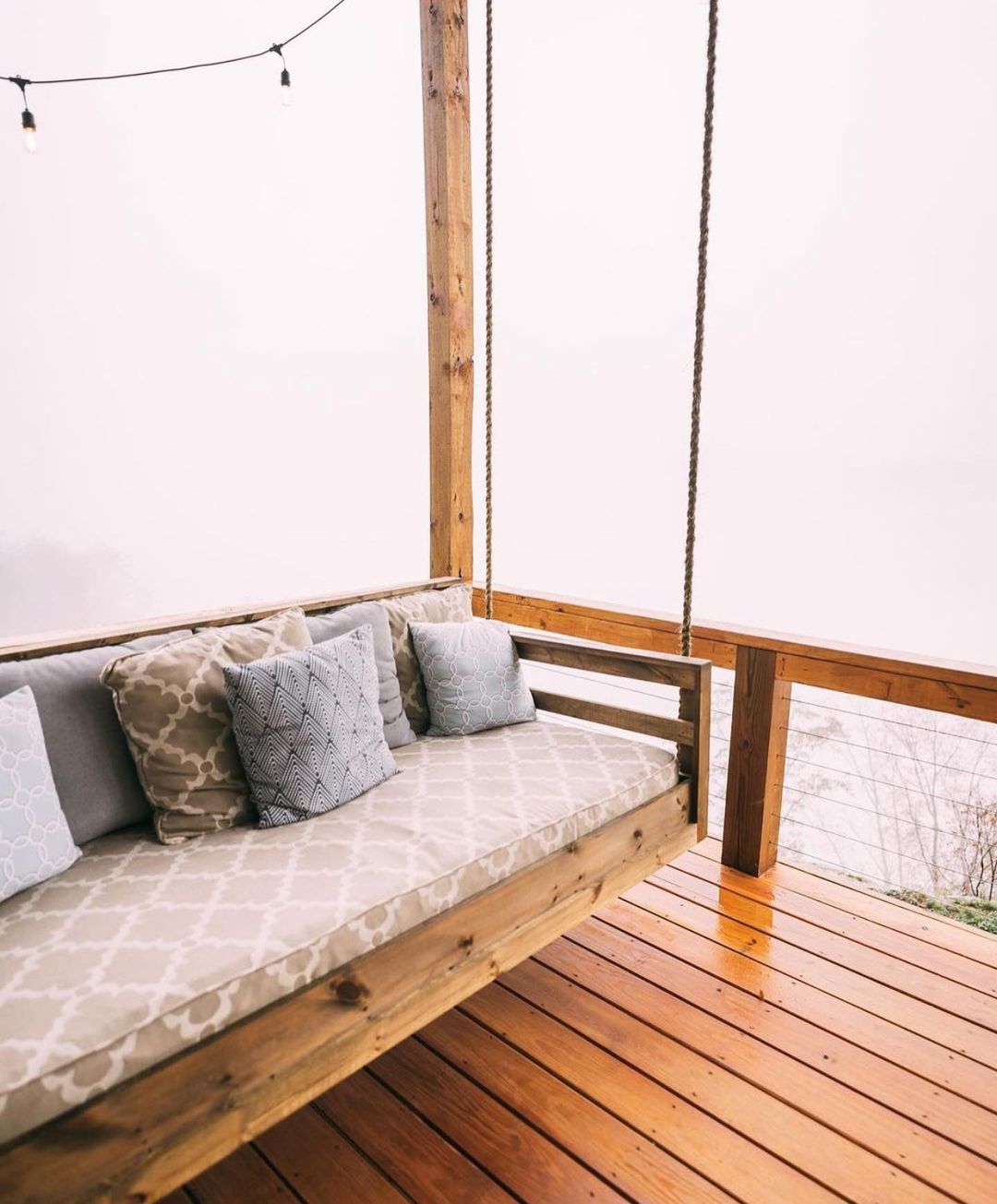 Wooden bench on the porch with a lot of pillows, and fairy lights above it