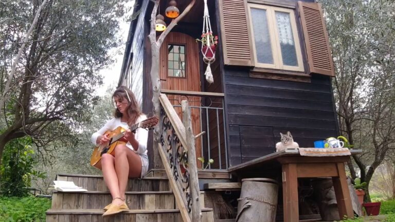 woman sitting on the stairs in front of her tiny house, playing guitar