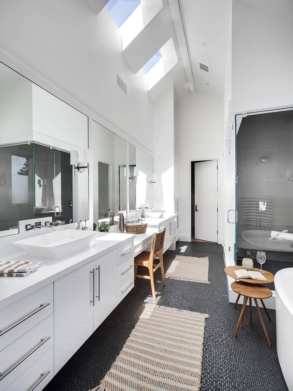 bathroom with a white bath, a glass shower with dark gray tiles, two sinks, two white cabinets and a long mirror