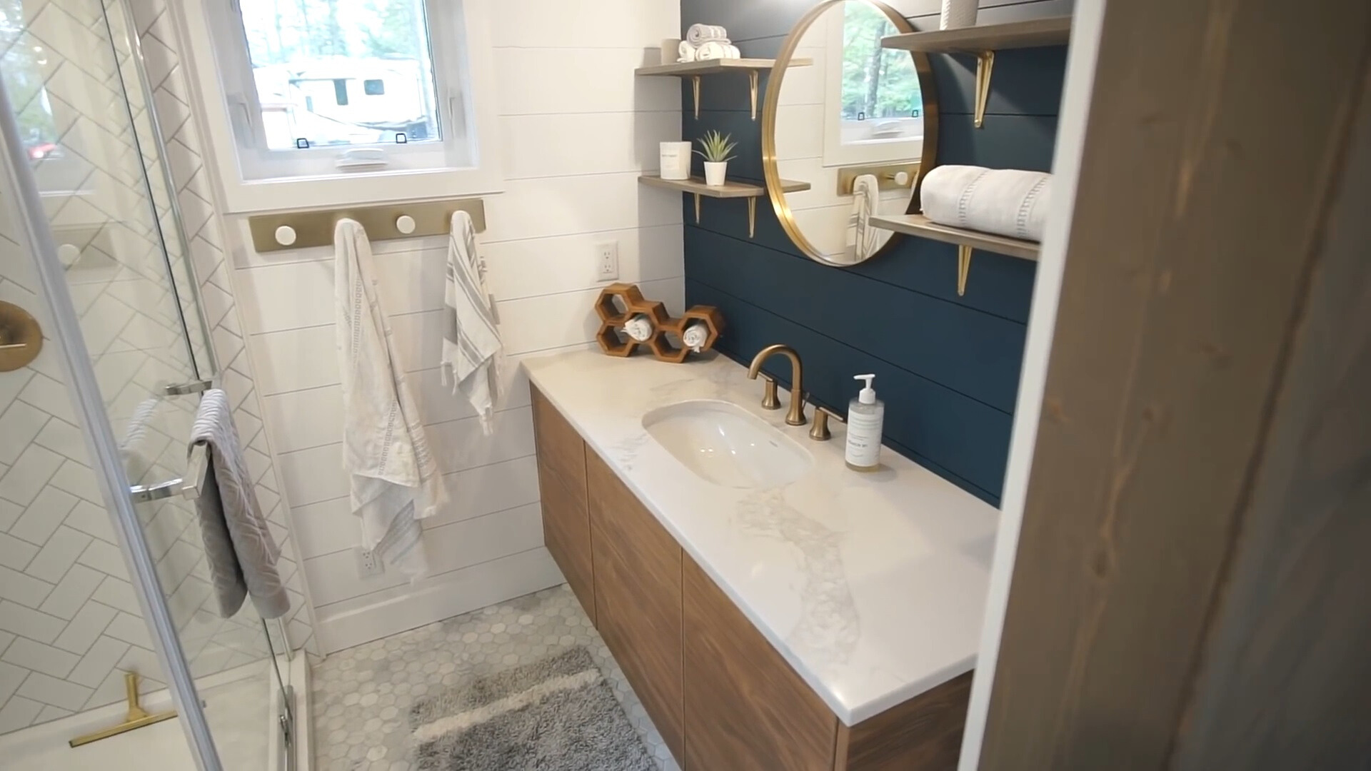 bathroom with white and dark blue tiles, glass shower, brown cabinet with a white countertop, shelves and a round mirror