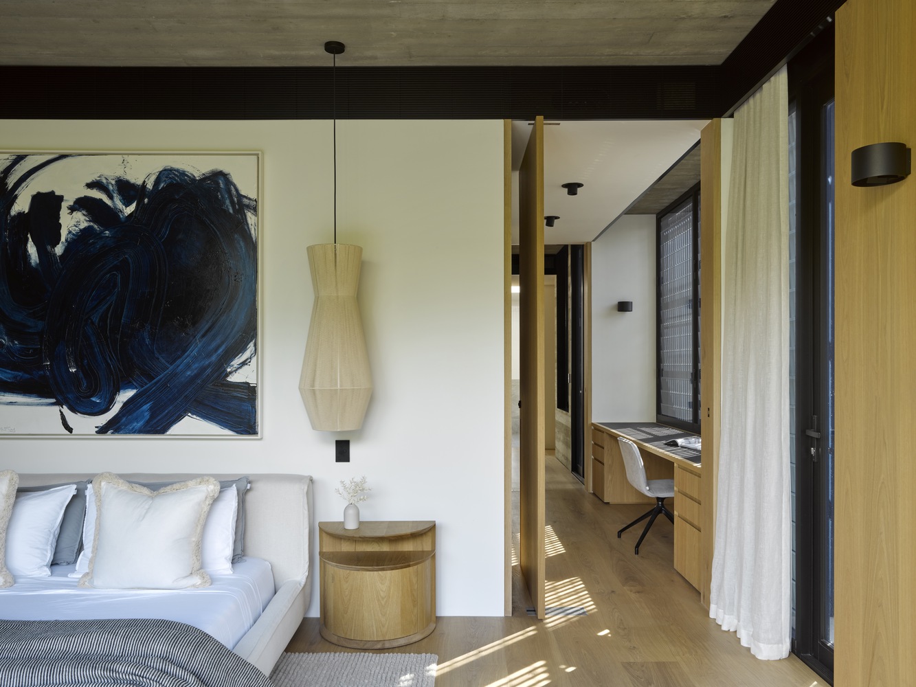 bedroom with a white bed, light colored bedsheets, blue modern art piece, brown bedside table, and an office area next to the room with a desk and a white chair