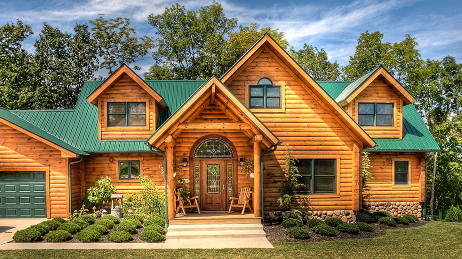 front side of the rustic log home the clear fork with orange-tint wood logs, a green roof, a garage, and greenery outside