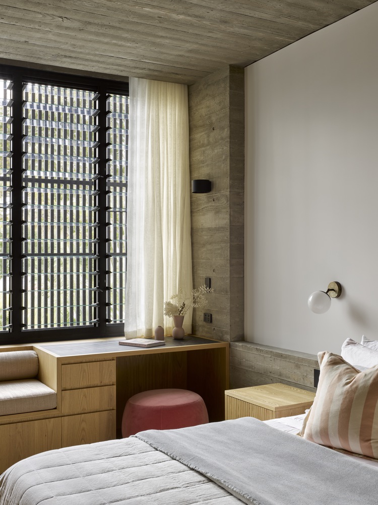 bedroom with white bed sheets, desk under a big table, a red stool and a brown bedside table