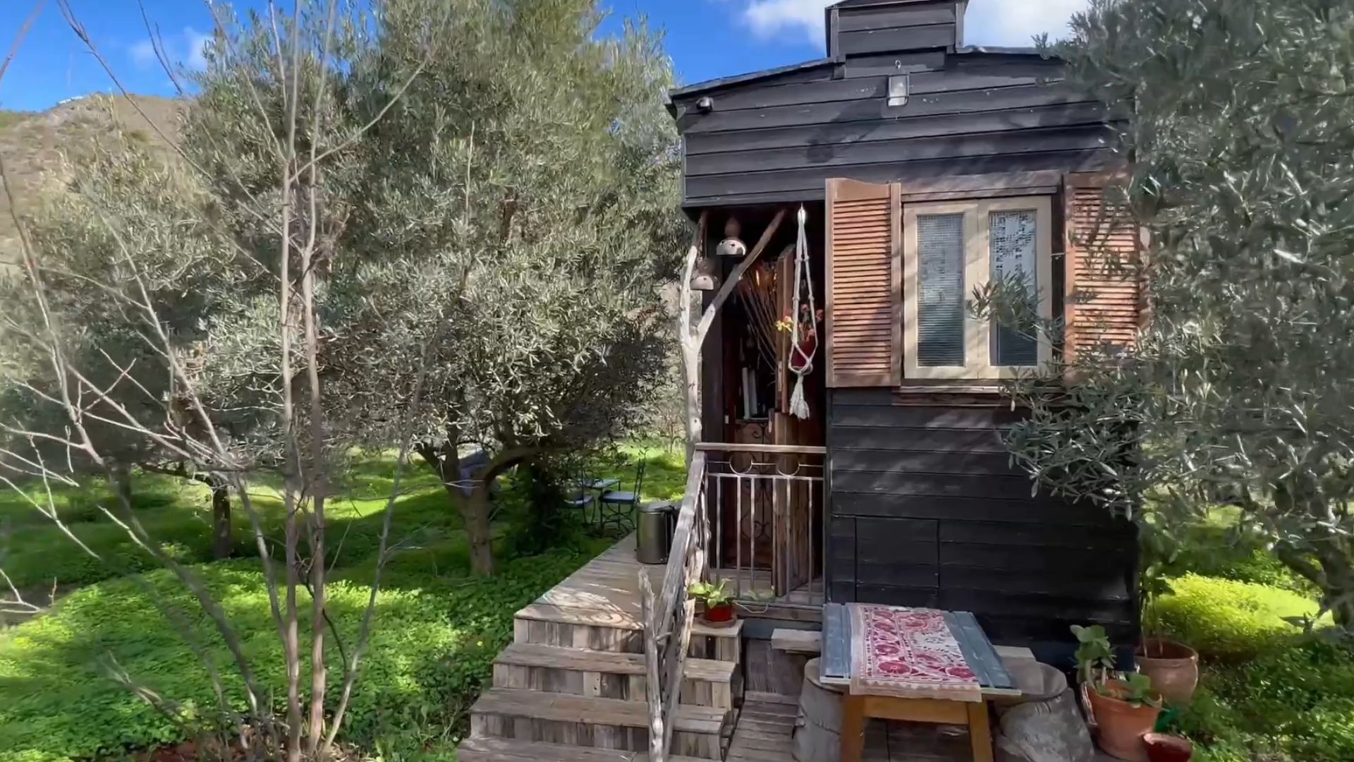 exxterior of the tiny house surrounded by olive trees, made using recycle materials, with a small sitting area in the front of the house