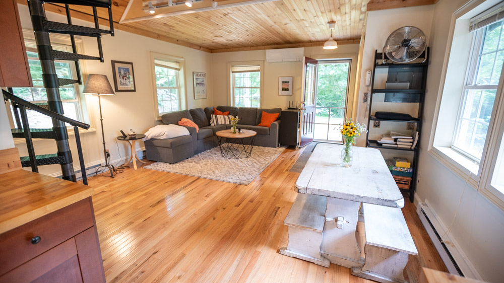 living room with a gray l-shaped couch, round brown table and a white painted wooden dining table with benches