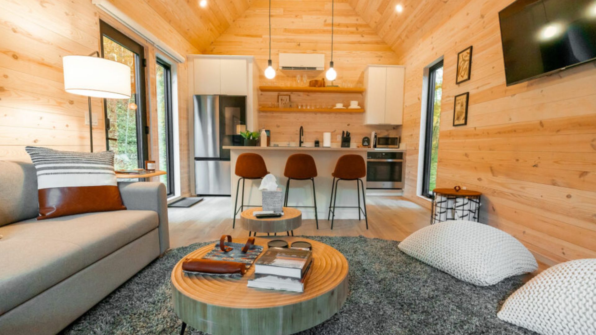 comfy living room with a light gray sofa, a round wooden coffee table, and a modern kitchen in the background featuring a bar counter with three brown stools