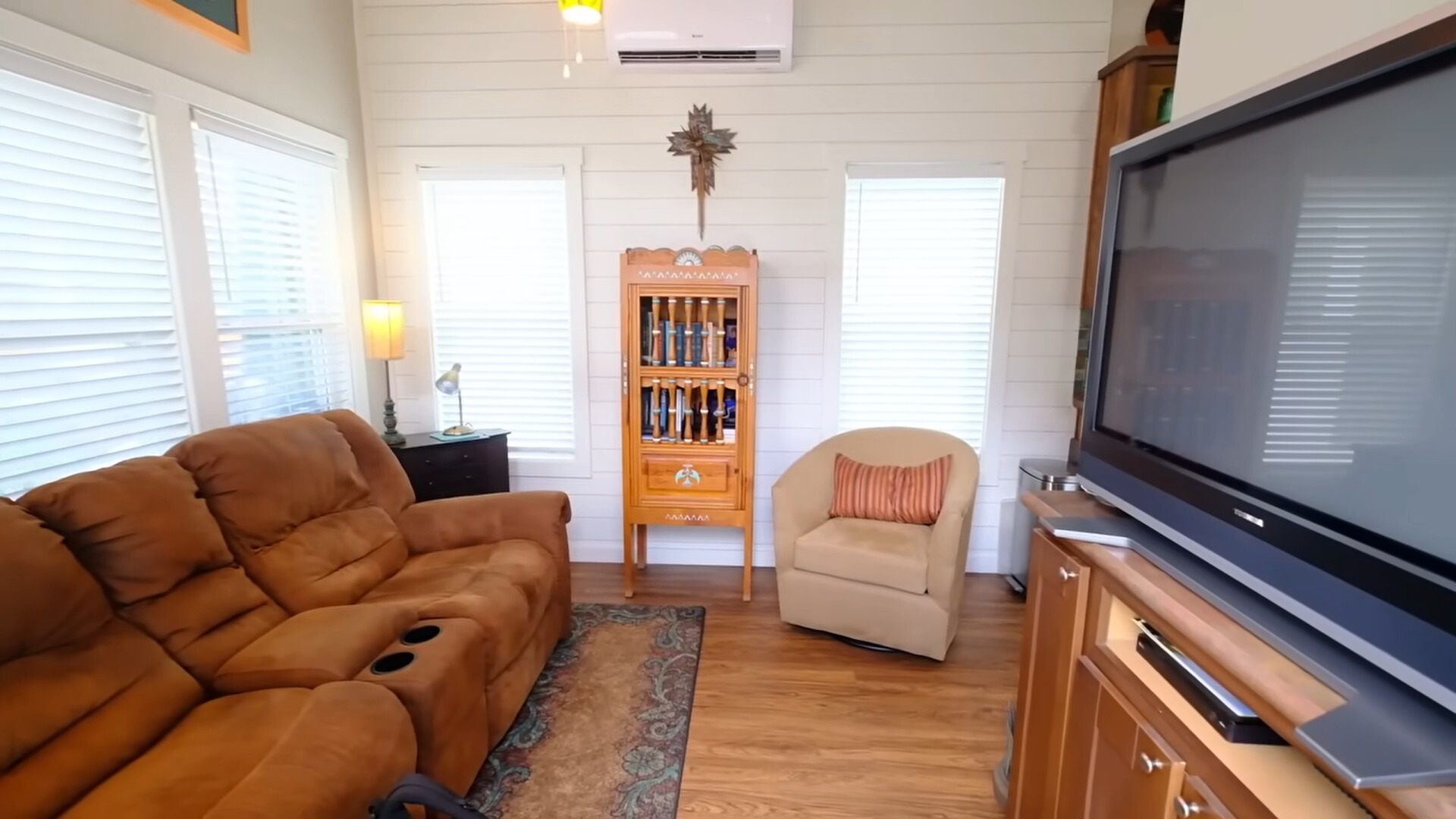 living room of a tiny house with a brown couch, a flat screen tv, cup holders inside a couch and windows all around the room