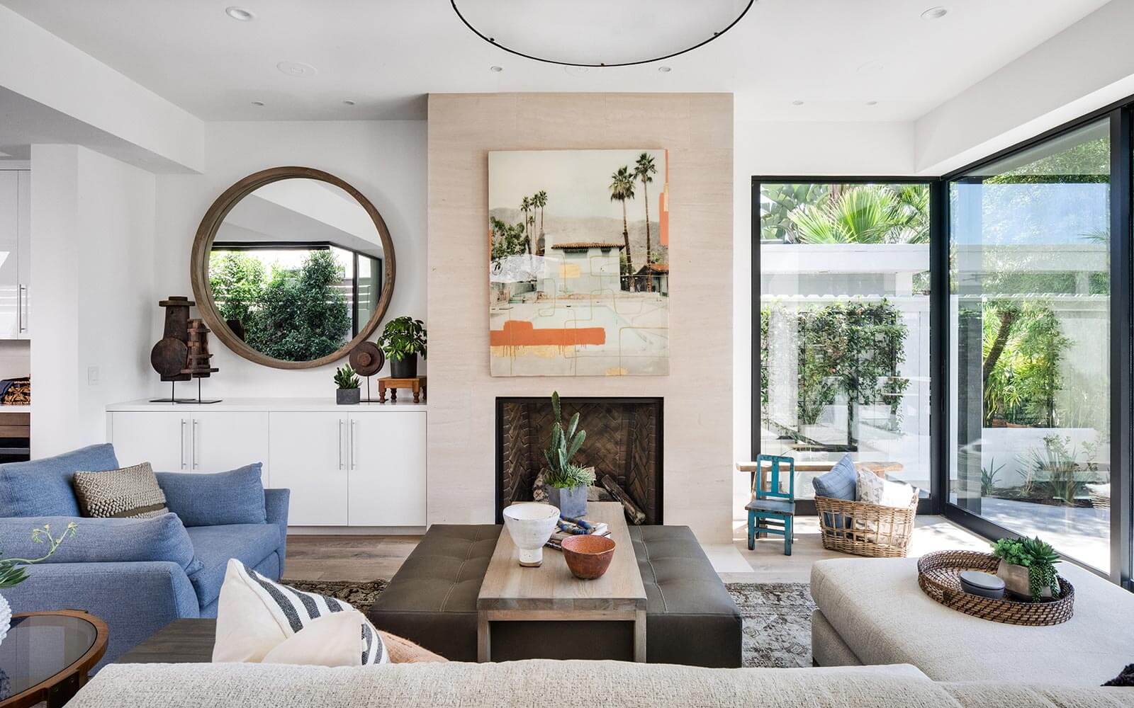 living room with floor to ceiling windows, big, round mirror, gray couch with a bloa sofa and a white cabinet and a fireplace under the art piece