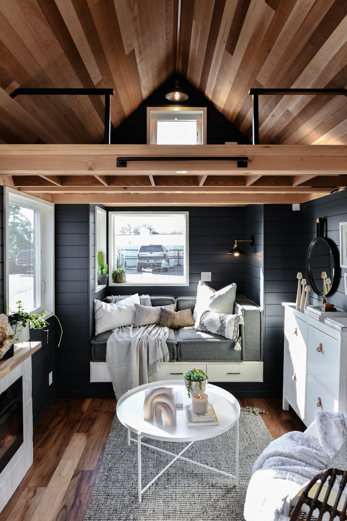 living room with a small gray couch, windows around it, dark walls, white table and a loft on top