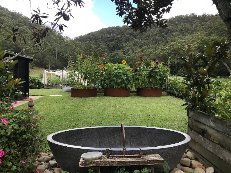 dark gray stone bathtub outside of the house, surrounded by flowers, fruits and plants