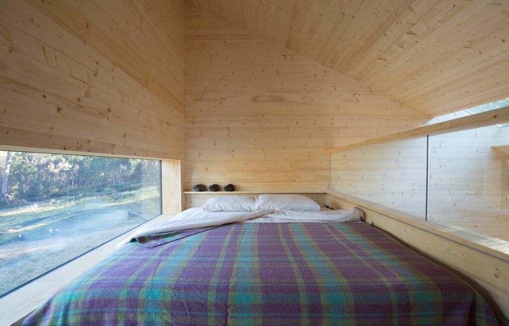 Full-sized bed in a wooden loft with a low window and a shelf