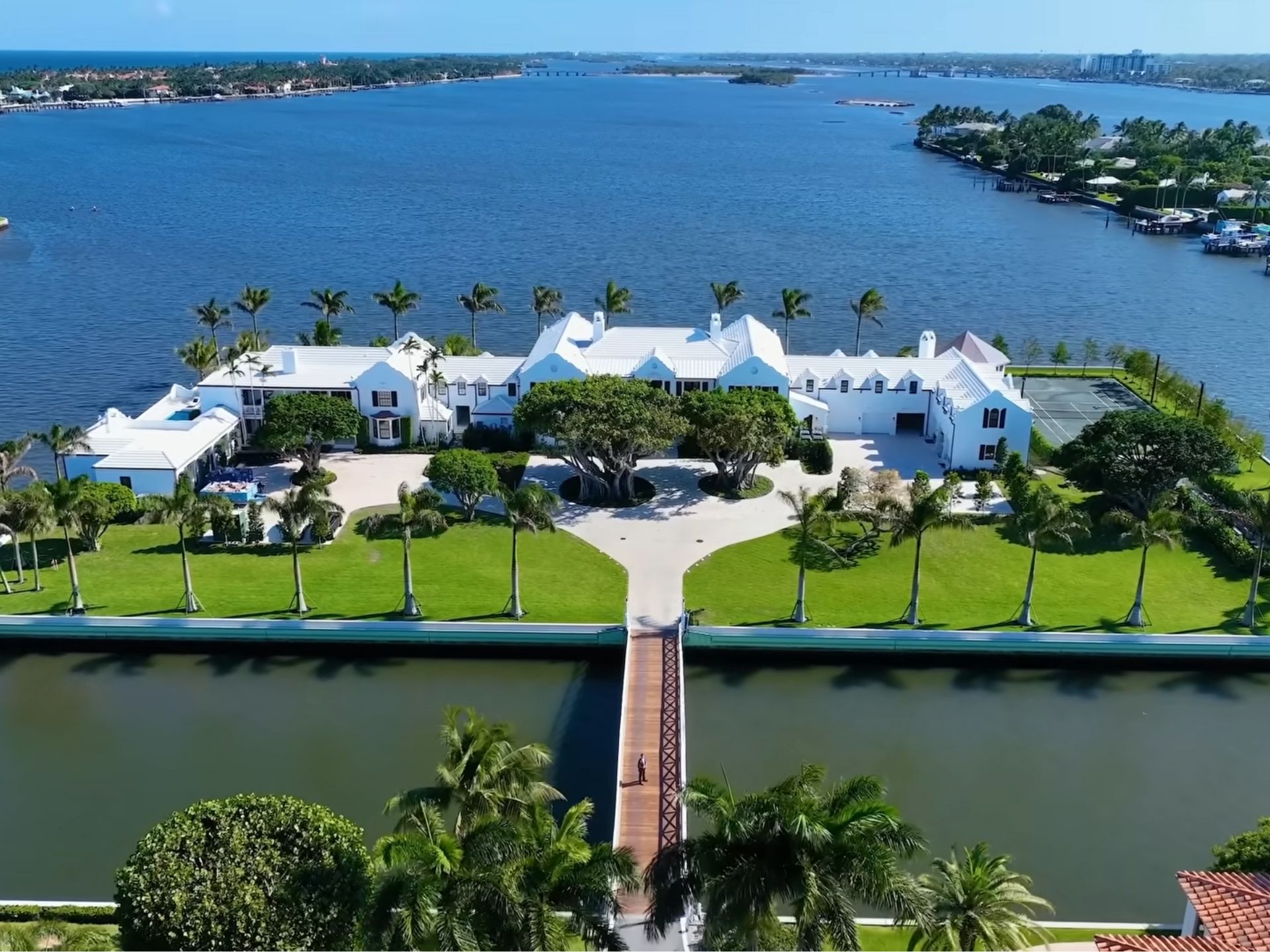 Aerial view of a luxurious waterfront estate featuring a large white mansion, lush green lawns, tall palm trees, a tennis court, and a bridge over a calm waterway, surrounded by water on all sides