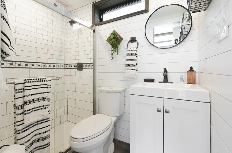 bathroom with a white cabinet, round mirror, hanging plant, big shower with glass doors and white tiles