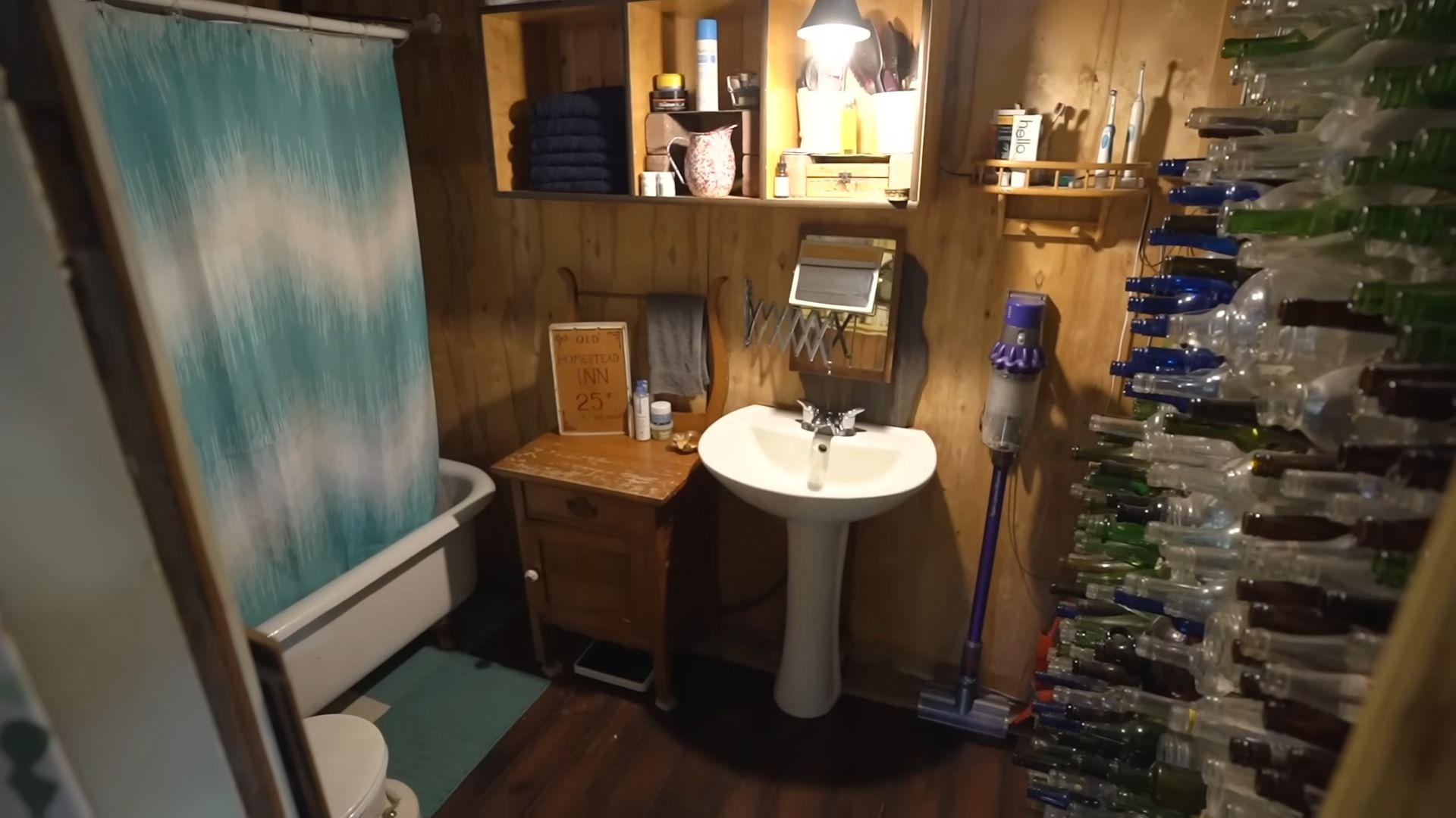 small bathroom with a tub and a blue curtain, a simple sink, compost toilet in the corner, and glass bottle wall on the right