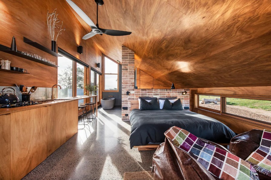 on the right is a bed with rectangular windows on the wall and on the left is a kitchen with brown cabinets and black shelves