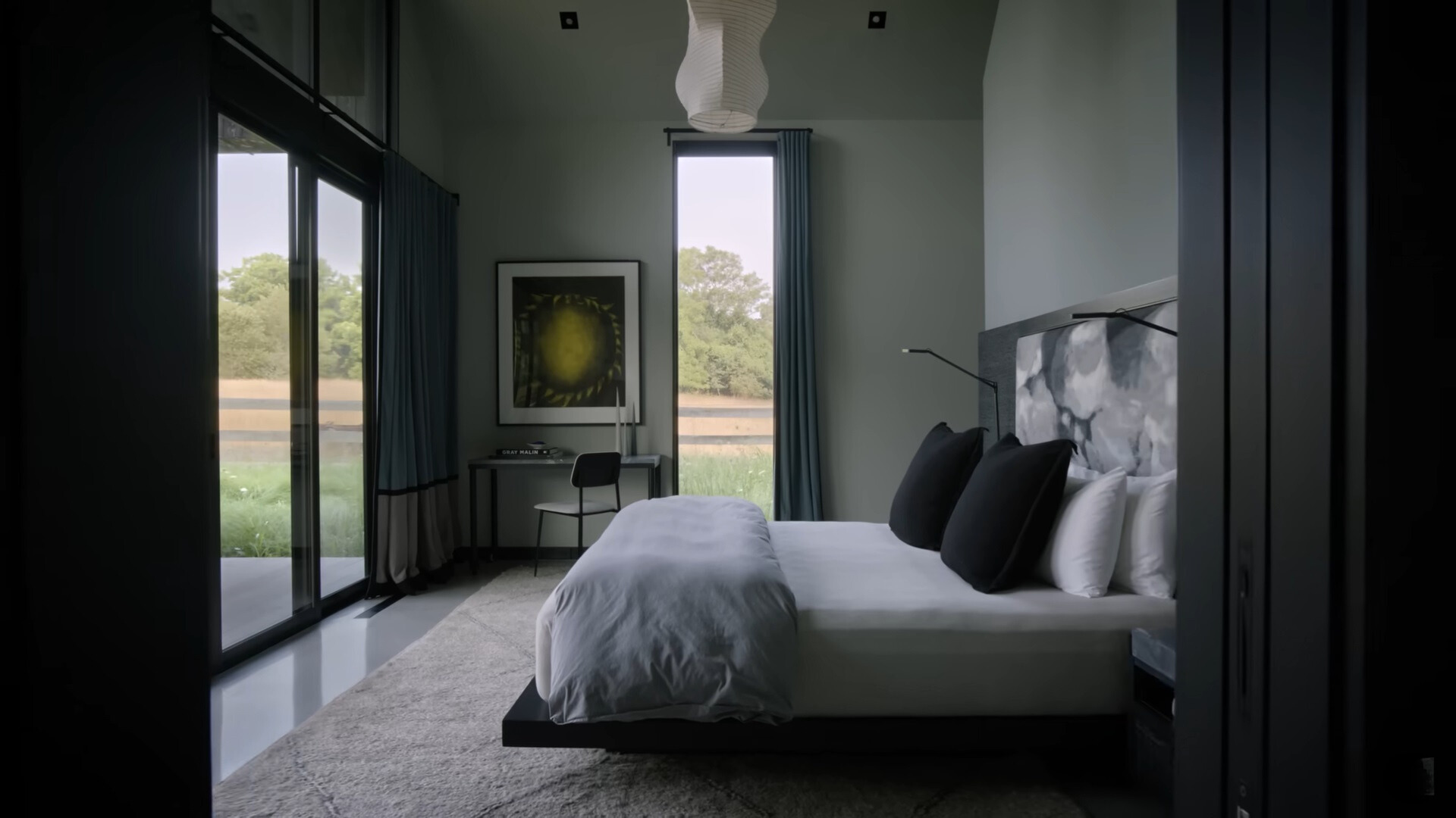 bedroom with gray walls, floor to ceiling windows, desk in the corner of the room with a small chair and a bed in the middle of the room with very simple lamps