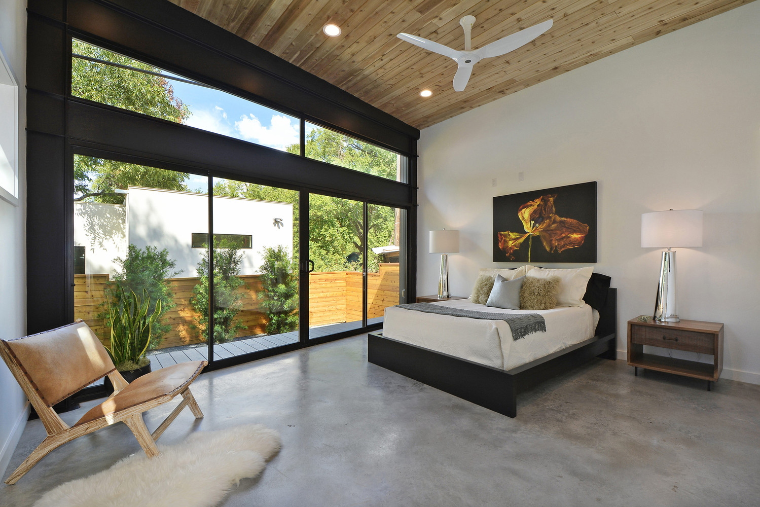 big bedroom with floor to ceiling windows, glass doors, a painting above the bed, bedside tables and a chair in the corner