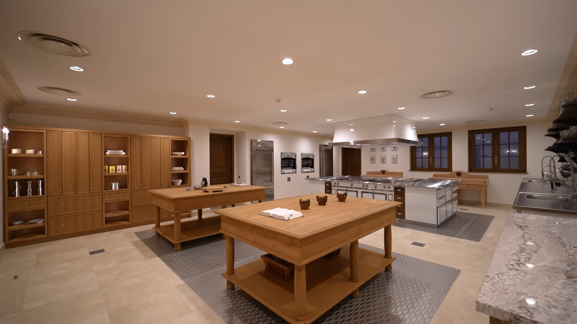 chef kitchen with two big brown tables, brown cabinets, ovens, fridge and sink