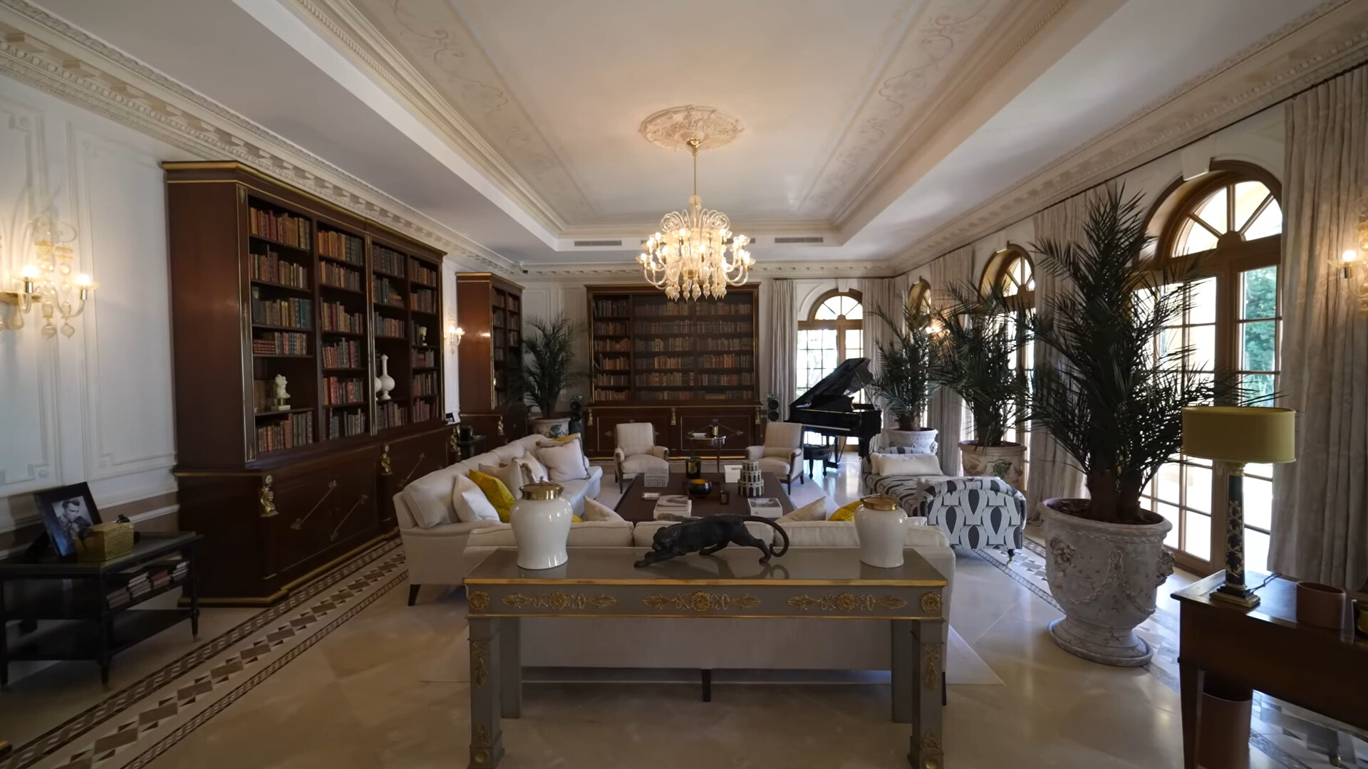 living room with floor to ceiling bookshelves on one side, big plants on the other and piano in the corner, an l-shaped white couch and big windows