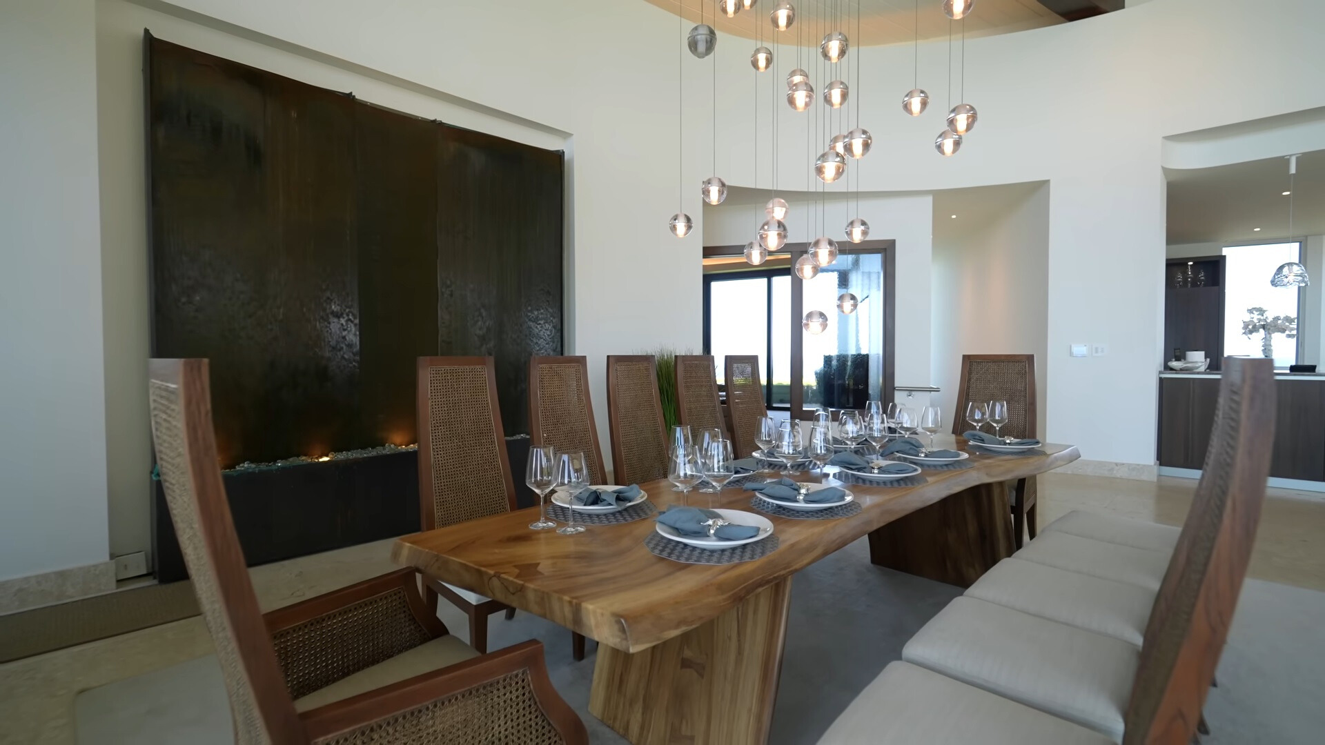 brown, wooden dining table surrounded by 12 chairs, low hanging chandelier and a waterfall wall