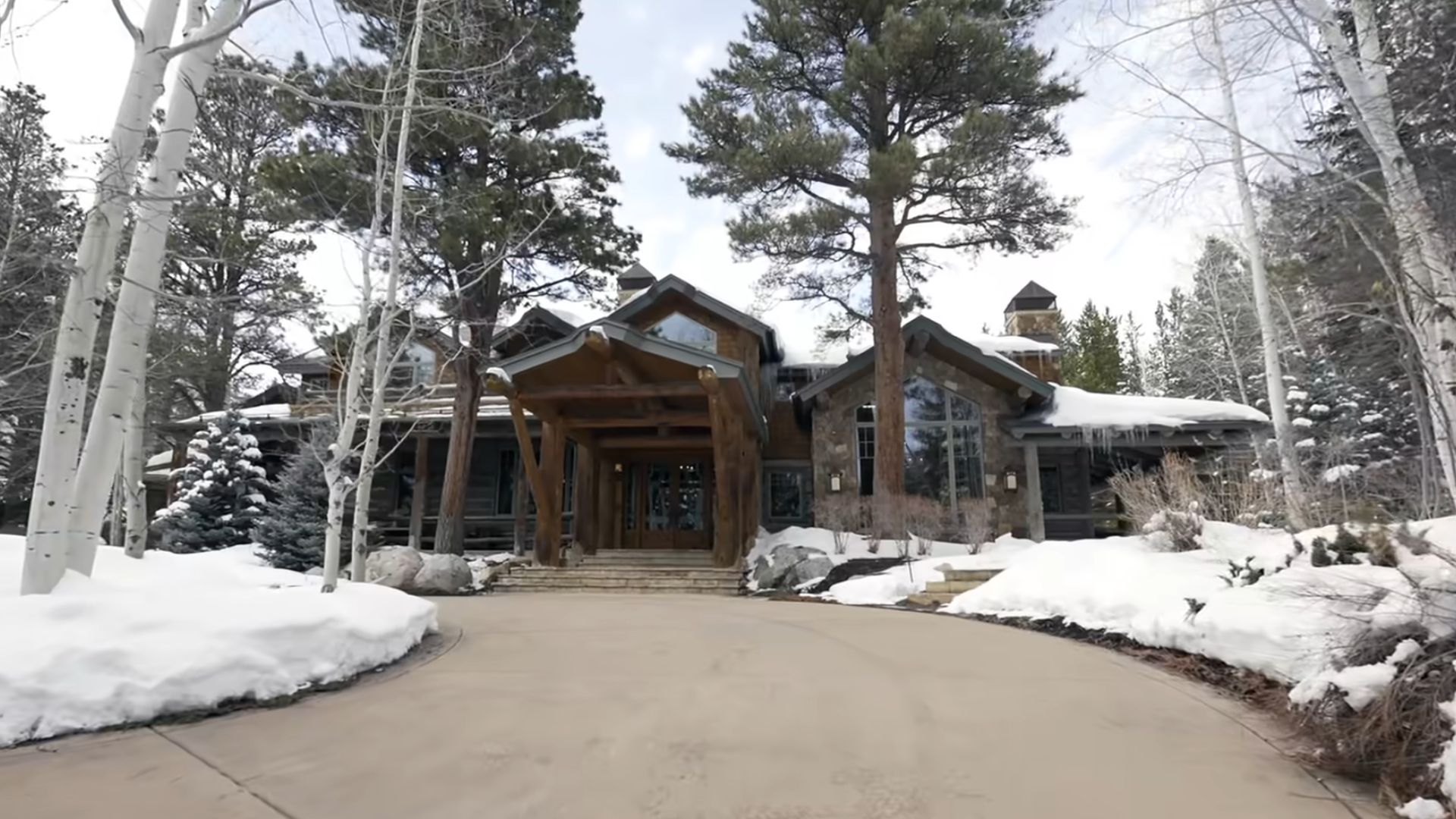 A large, rustic-style mountain cabin with a stone and wood exterior, surrounded by snow-covered trees and landscape, a steeply pitched roof, large windows, and a welcoming entrance with a wooden pergola