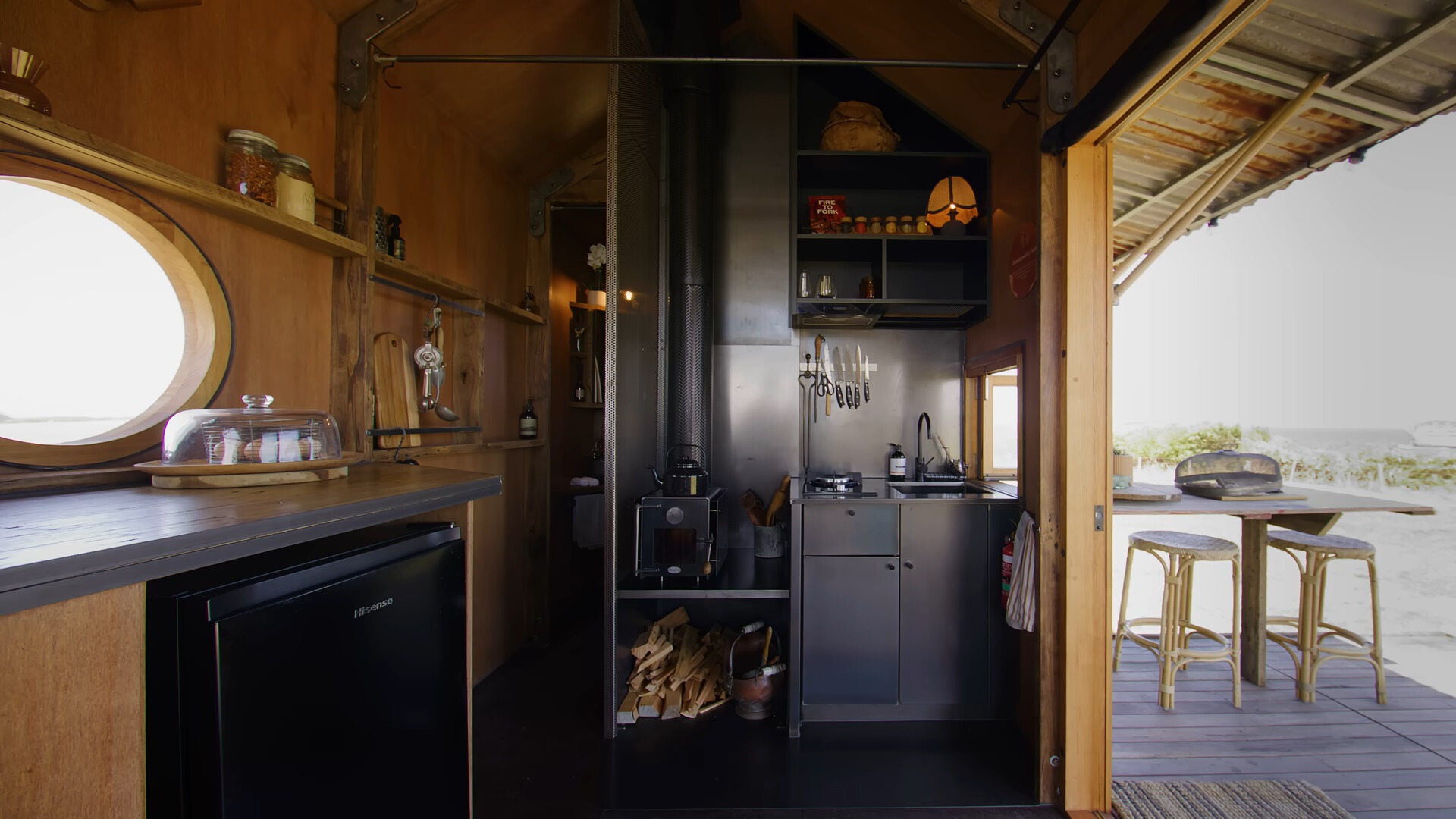 kitchen of a tiny home with a countertop on the left with shelves above it and one more small countertop with a sink and black cabinets, on the outside is a dining table with bar stools