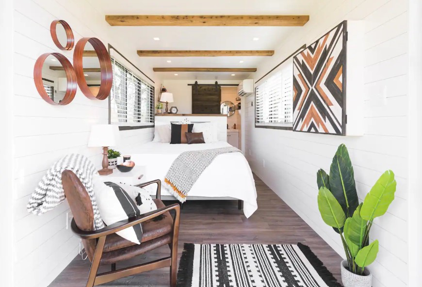 living room and bedroom of a container house with a comfy bed, two windows on each side of the container, a brown chair in the corner of the room next to a white cabinet with a tiny lamp and round mirrors of different sizes