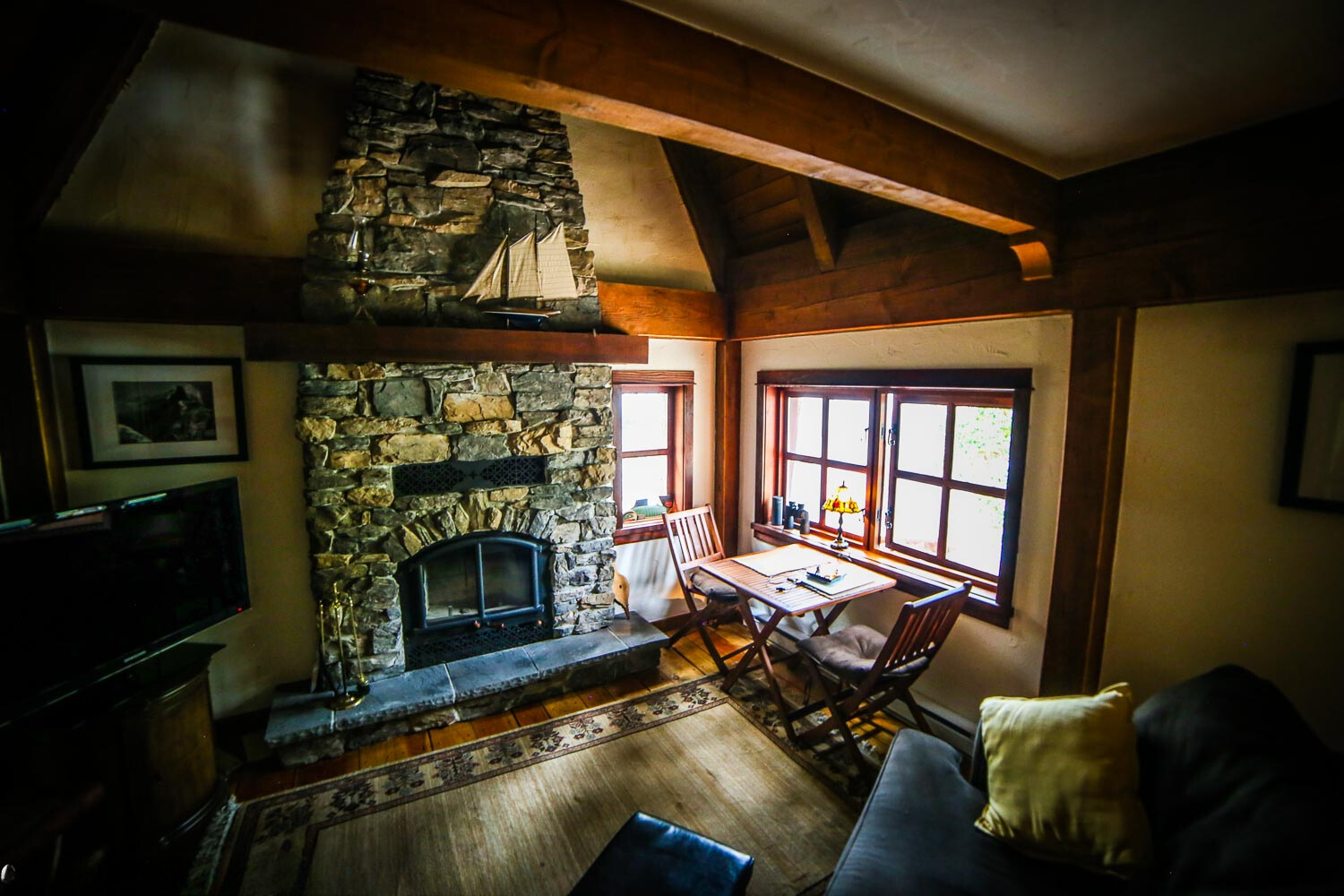 living room with a flat screen tv, small brown table next to the window and a fireplace surrounded by stone