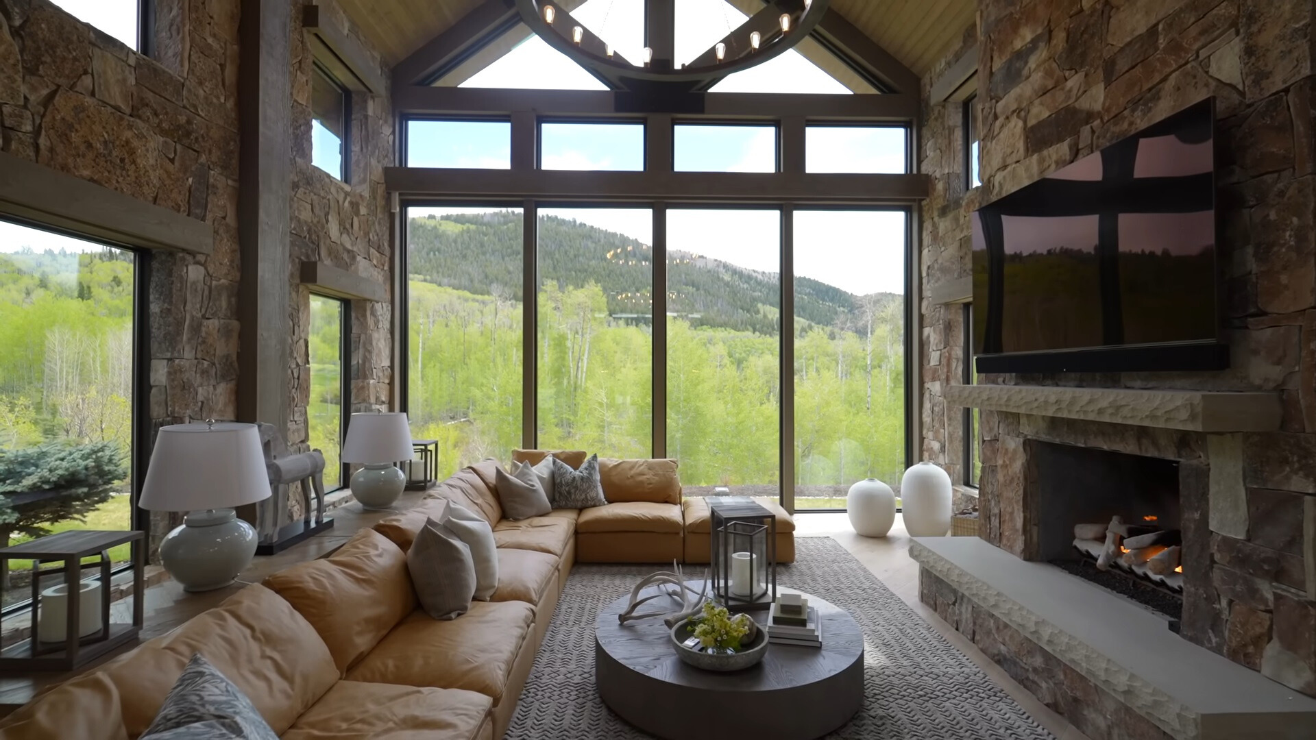 living room of a big cabin with a floor to ceiling windows looking onto woods, orange couch, flat screen tv, round table and a fireplace