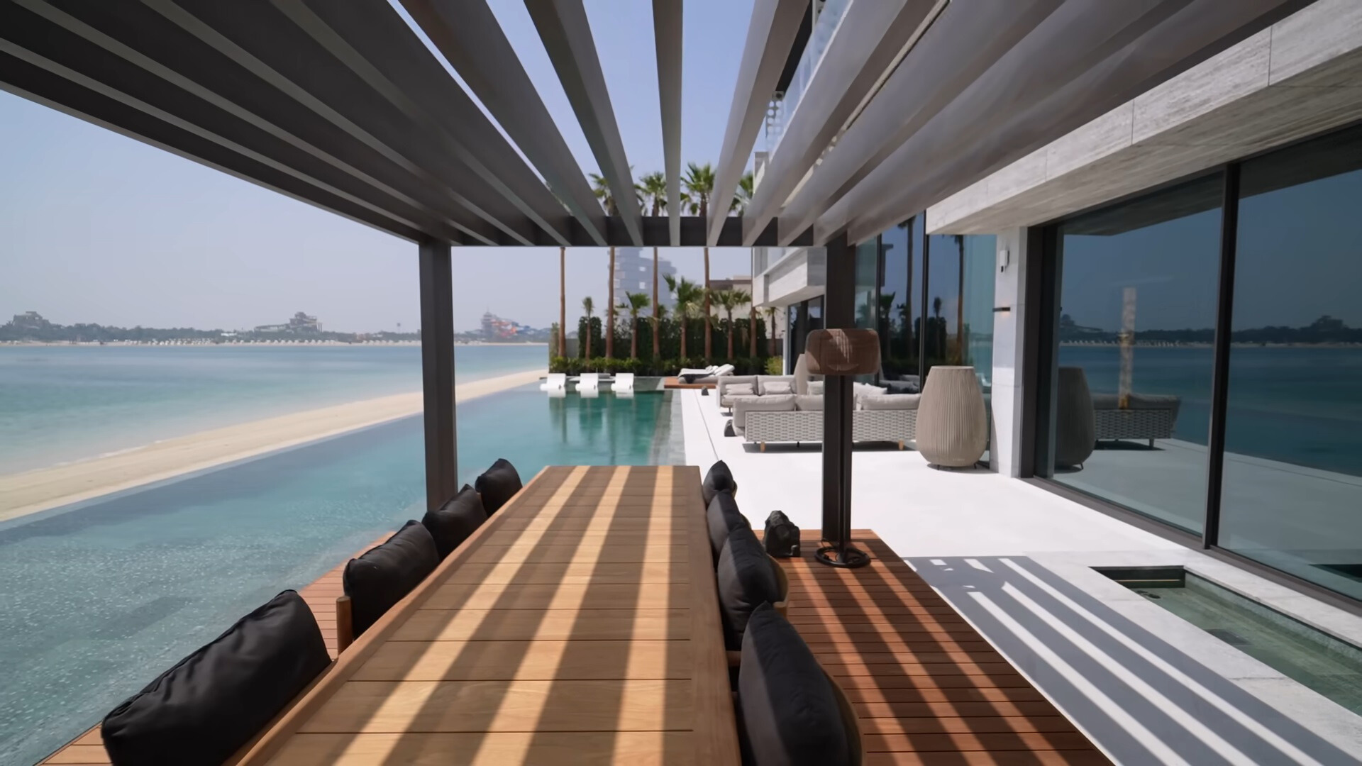 outdoors area of a modern house with a long pool, sitting area and a dining table with chairs that's covered