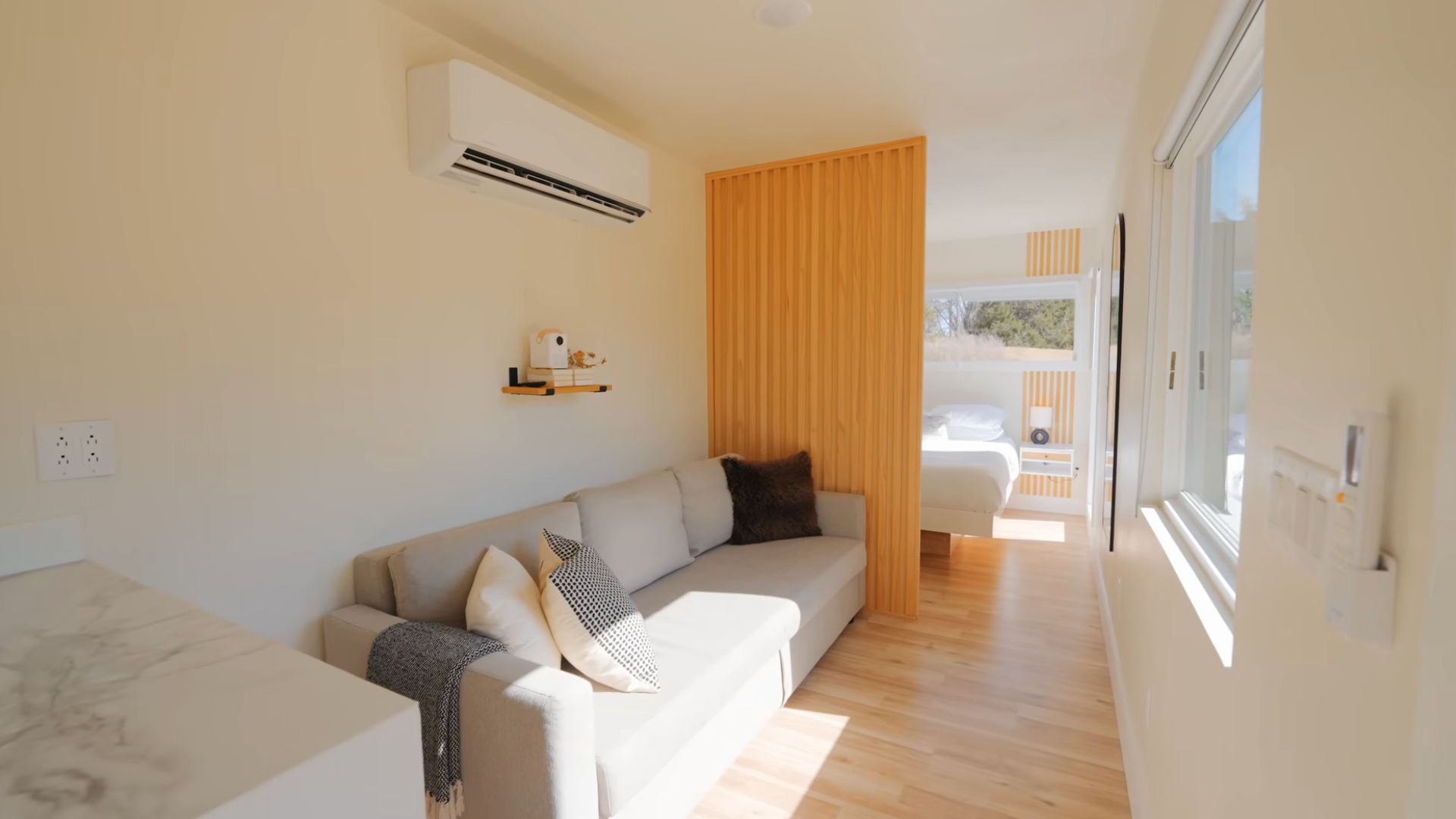 living room area with a light-colored couch, AC on the wall, nestled between the kitchenette and the bedroom, right next to the wooden panel divider