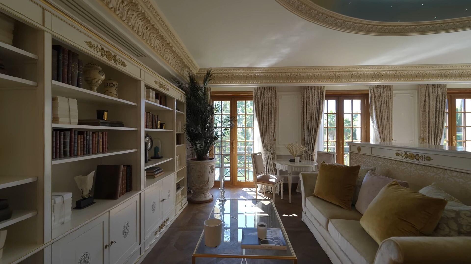 white couch in a living room, a glass table, round white table with chairs in the background, opposite the couch a big shelf with books and other decor