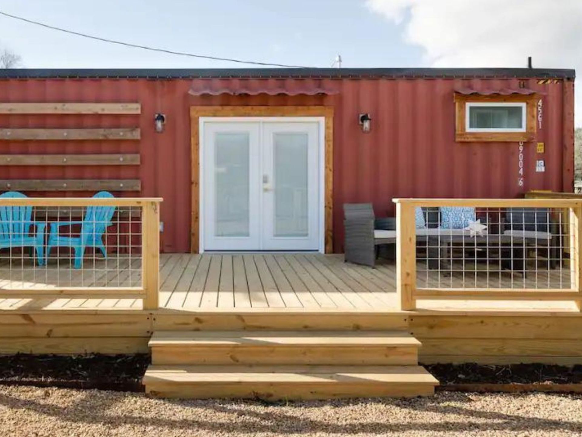 exterior of the container house with red corrugated siding, a spacious deck with a wire fence, two seating aareas, some wooden shelves on the left side of the container near the entrance