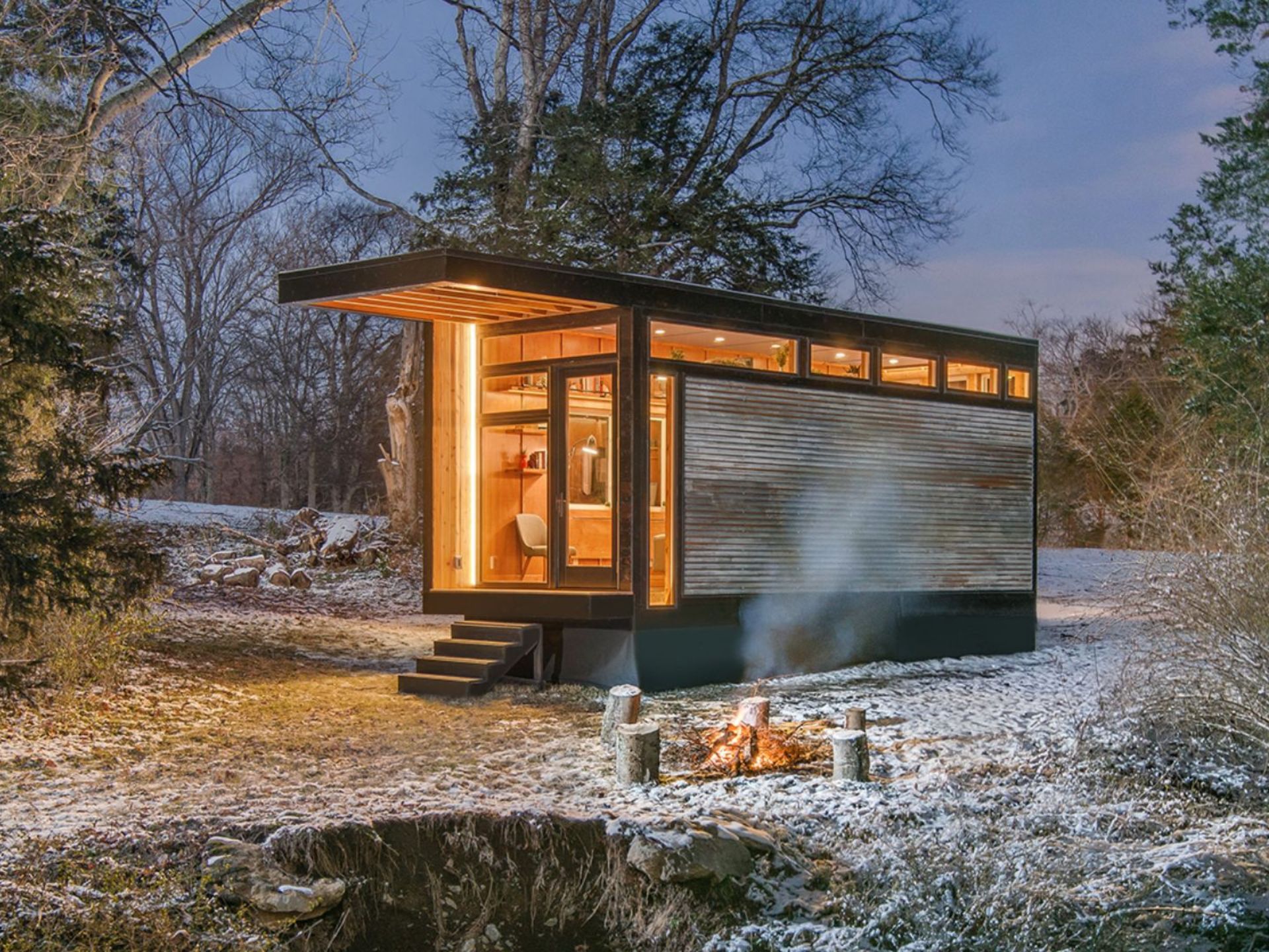 exterior of the tiny house Cornelia with wood siding, black steel frame, small stairs, lights on at night