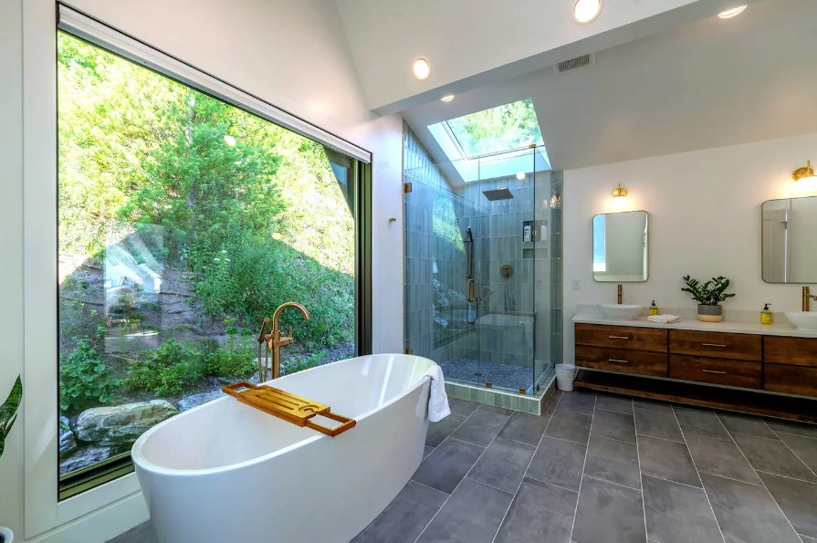 bathroom with a white bathtub next to a huge window and a glass shower with blue tiles and a ceiling window above