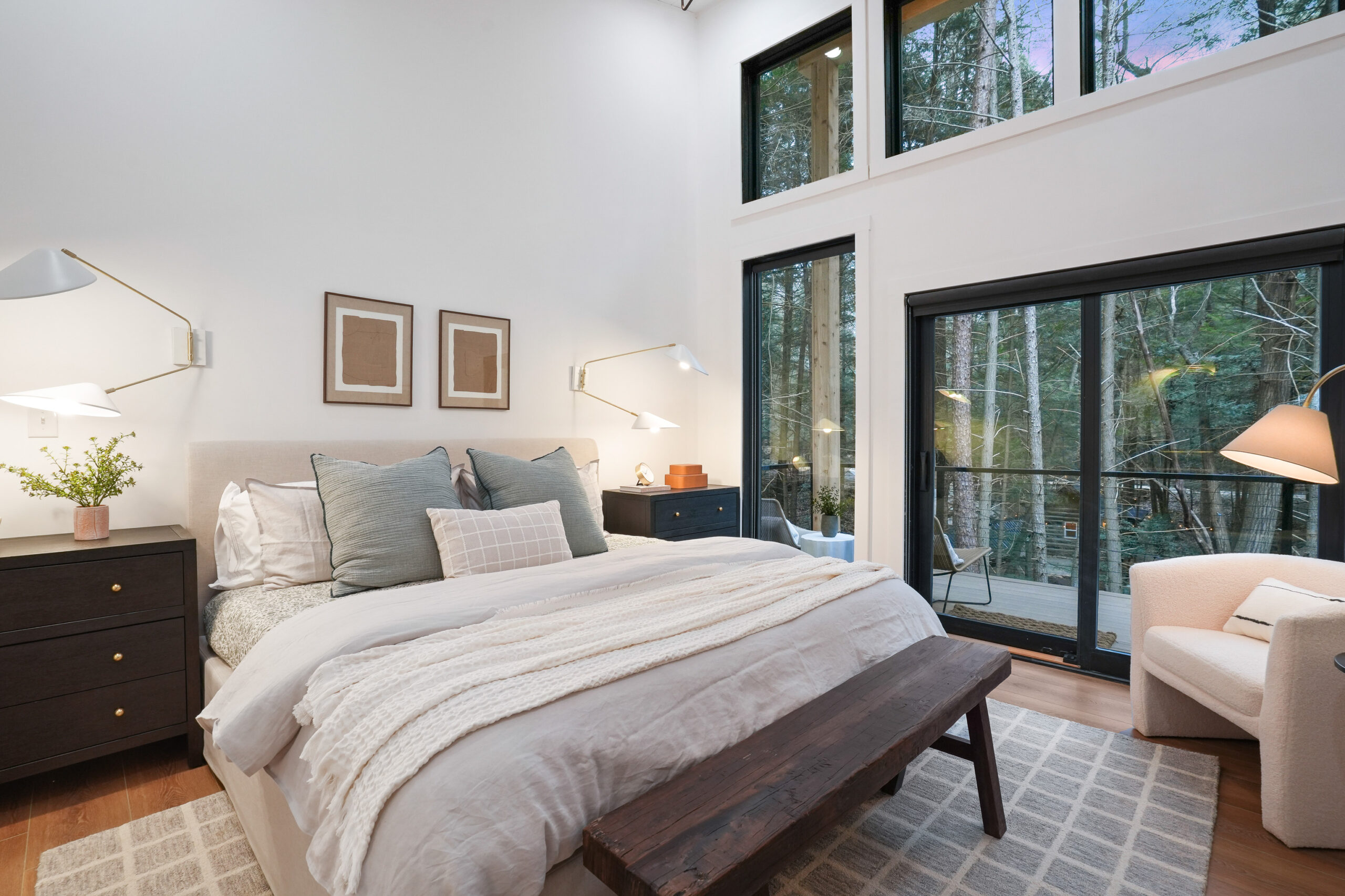 bedroom with a king sized bed, big windows with an opening to a deck, dark brown bedside tables with lamps, white sofa in the corner