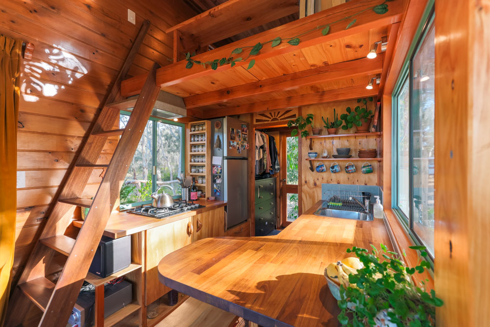 kitchen of a tiny house with wooden countertops, wooden walls, open shelves, big windows above the countertops