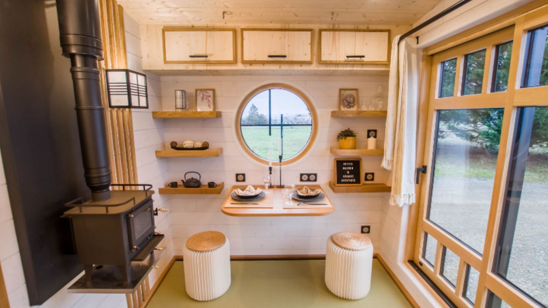 a cozy, compact living area with a wood stove, a circular window, and a small dining setup with stools in a wooden tiny house