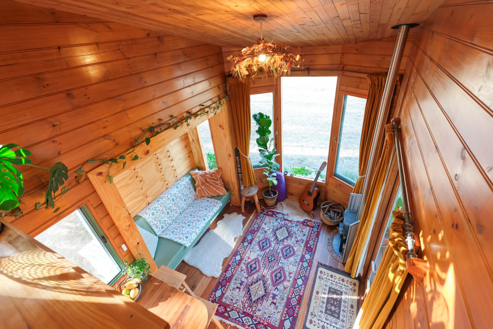 living room of a tiny house with a green couch, patterned carpet, wooden walls, big windows, plants