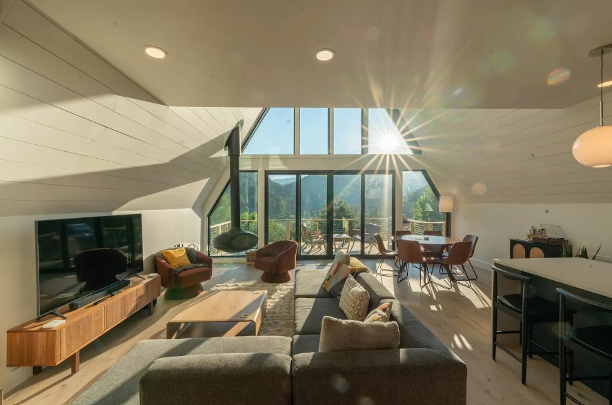 living room with an l-shaped couch, floor to ceiling windows, flat-screen tv on the brown cabinet, hanging fireplace with brown chairs and a round brown table with chairs around it