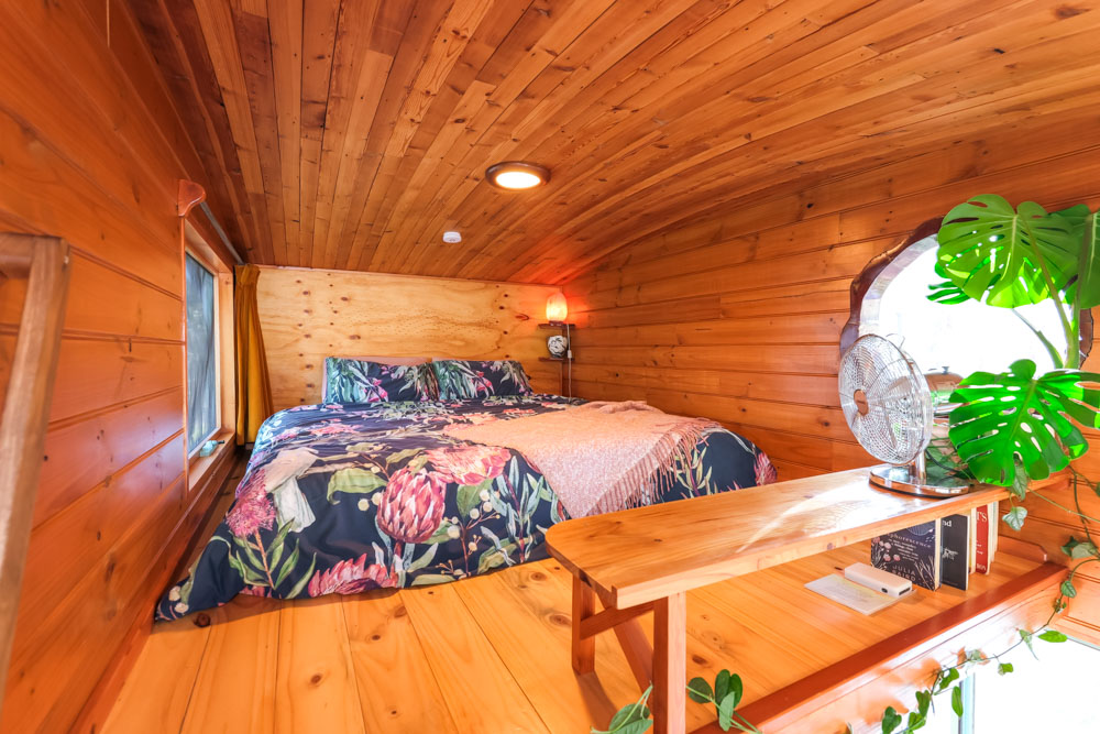 loft bedroom with wooden walls, floral bed sheets, round window and a mini table which is also a shelf