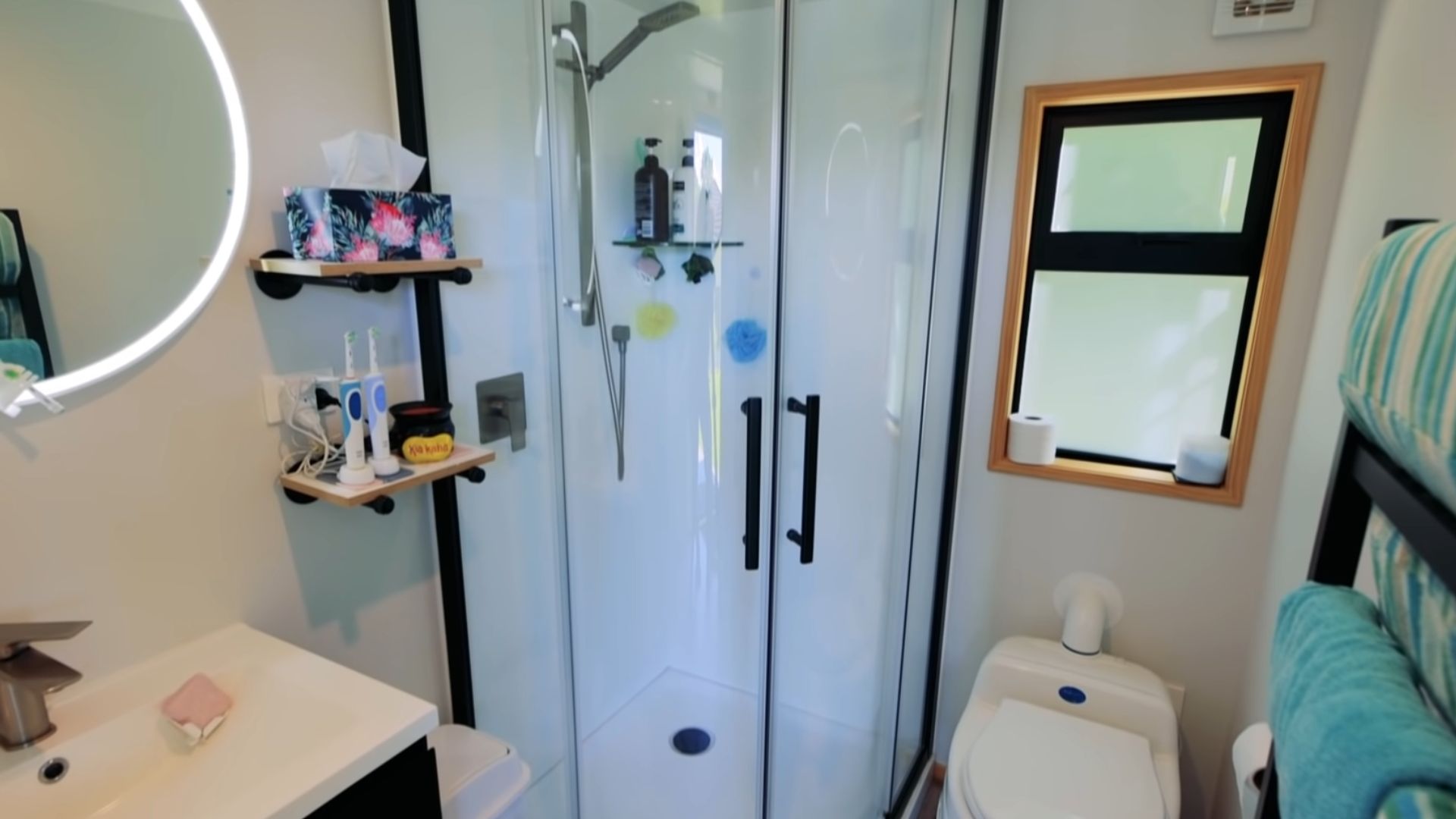 small bathroom in the tiny house with a shower, toilet, sink, and a light-up round mirror, black hardware on the shower