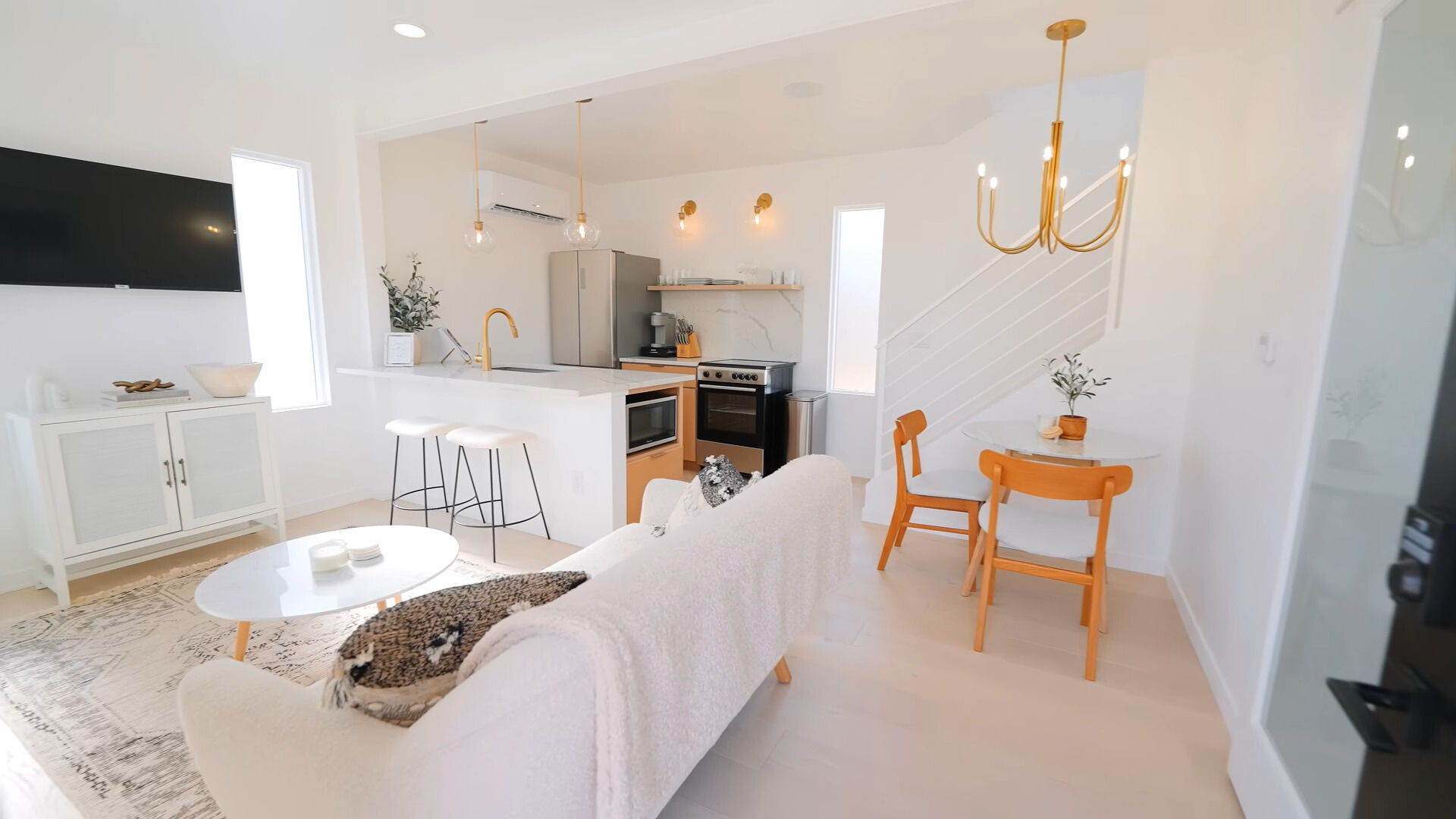 white couch of a living room, white oval table, white cabinet under a flat screen tv, white island with two bar stools in the kitchen, white round table in the corner accompanied by two chairs