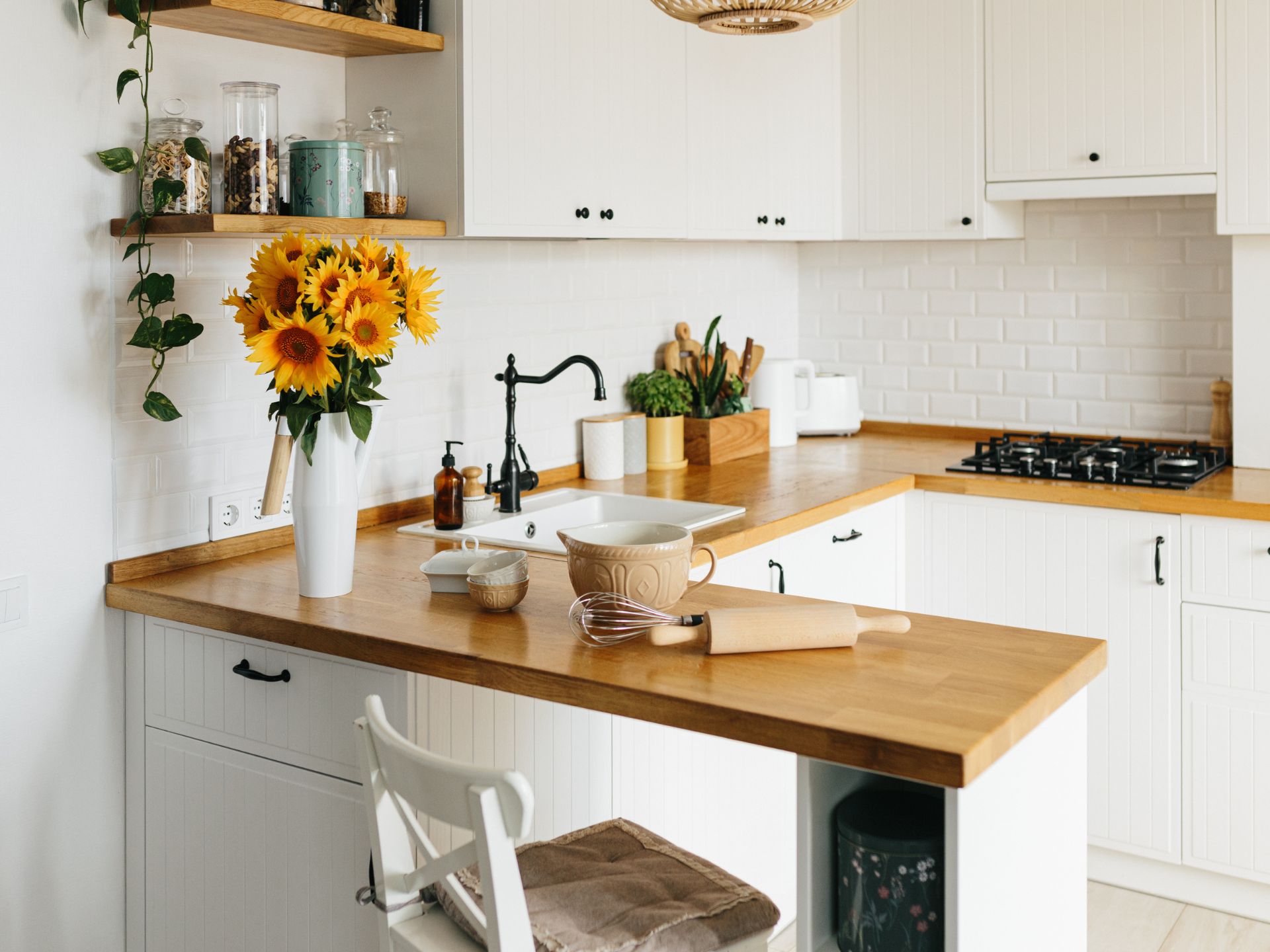 small L-shaped white kitchen with beadboard cabinets, wooden countertop, white metro tiles as baksplash, two open shelves on the left, a vase full of sunflowers on the counter