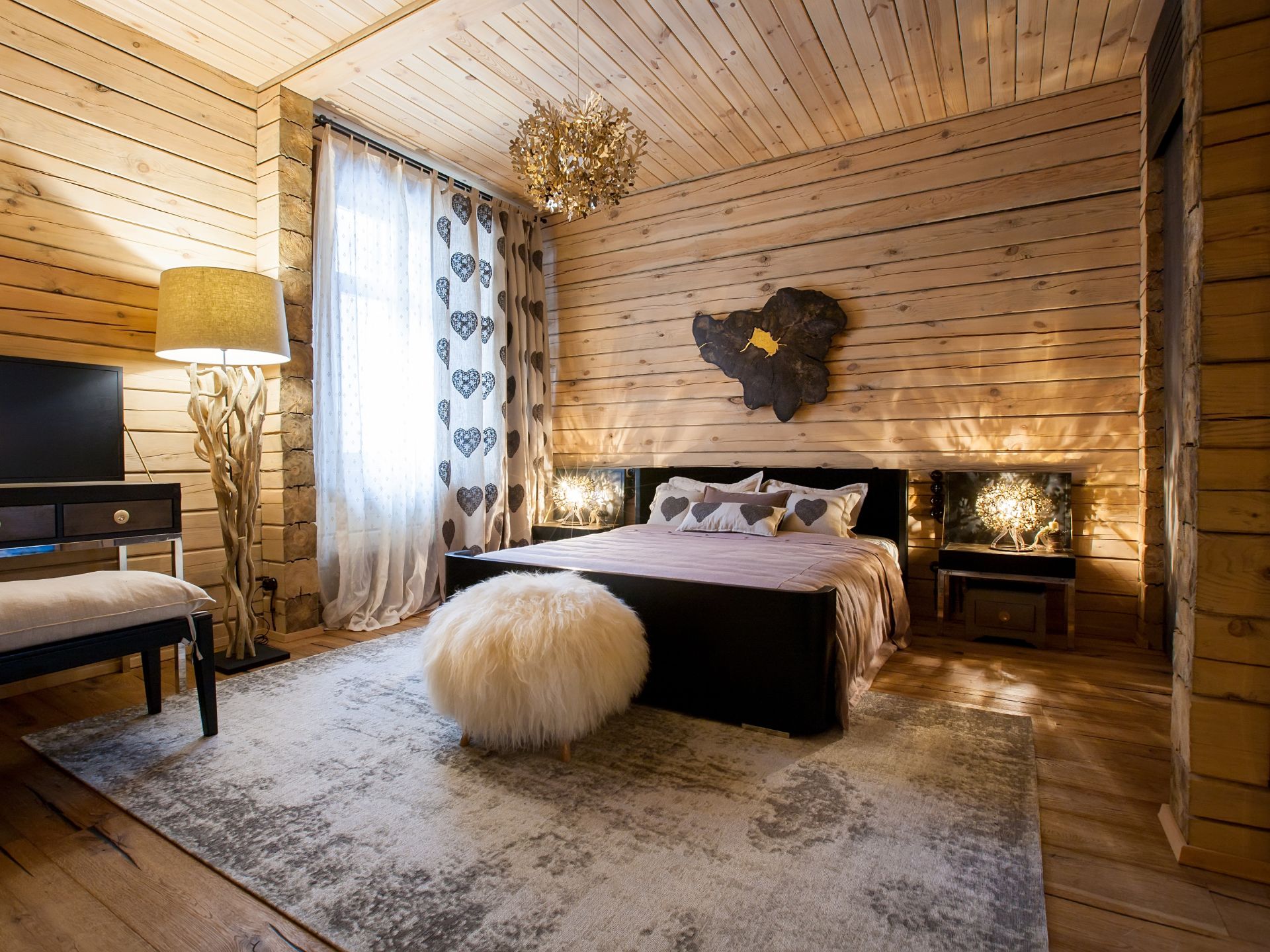 rustic bedroom with wooden wall siding, a classic bed in the center, a cool art piece above it in black and gold, a faux fur ottoman chair, a golden chandelier, and a makeup table on the left