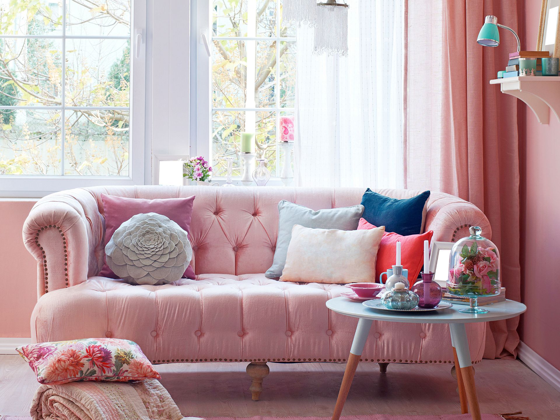 shabby chic living room idea with a gorgeous, elegant pink sofa, lots of throwpillows, some floral ones on the floor, a small round side table in light blue with some candles and flower arrangements on it, windows in the bavk with pink curtains and pink walls