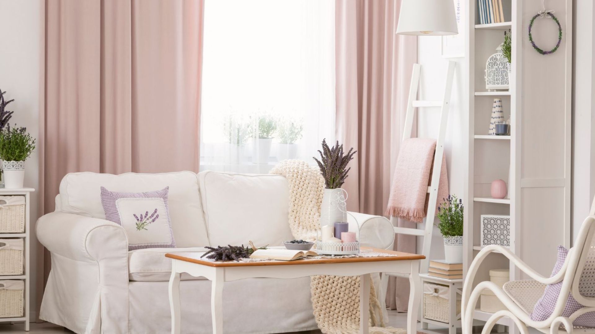 beige shabby chic living room with a simple coffee table, a ladder shelf next to it with a soft pink blanket, cubicle shelves, a white rocking chair, and beautiful blush pink curtains on the walls matching with the same pink rug