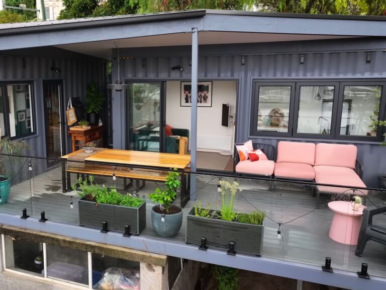 terrace of the container house with pink lounge area on the right, a large dining table in the middle of the terrace, two black containers with corrugated walls combined