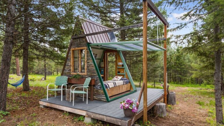 tiny a-frame cabin with a right side opening completely, bed inside, a small porch around the cabin and two chairs